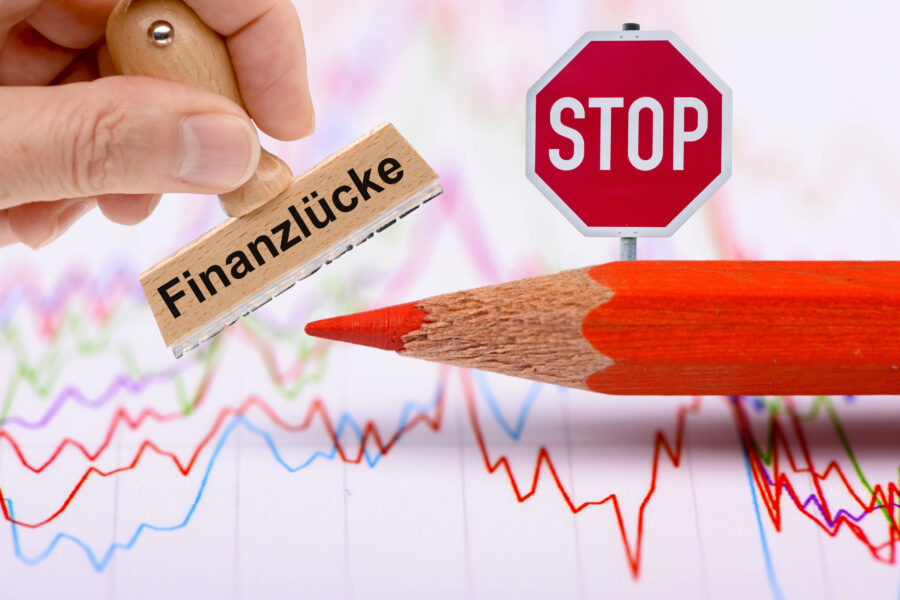 Roter Bleistift neben Stempel mit "Finanzlücke" und "STOP"-Schild, im Hintergrund Diagramme.