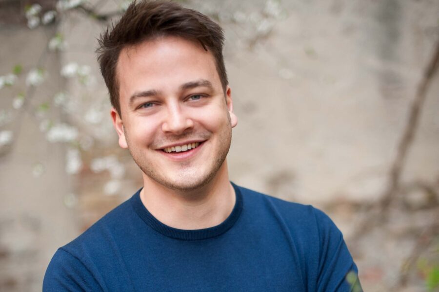 Eine lächelnde Person mit blauem Pullover vor unscharfem Hintergrund.