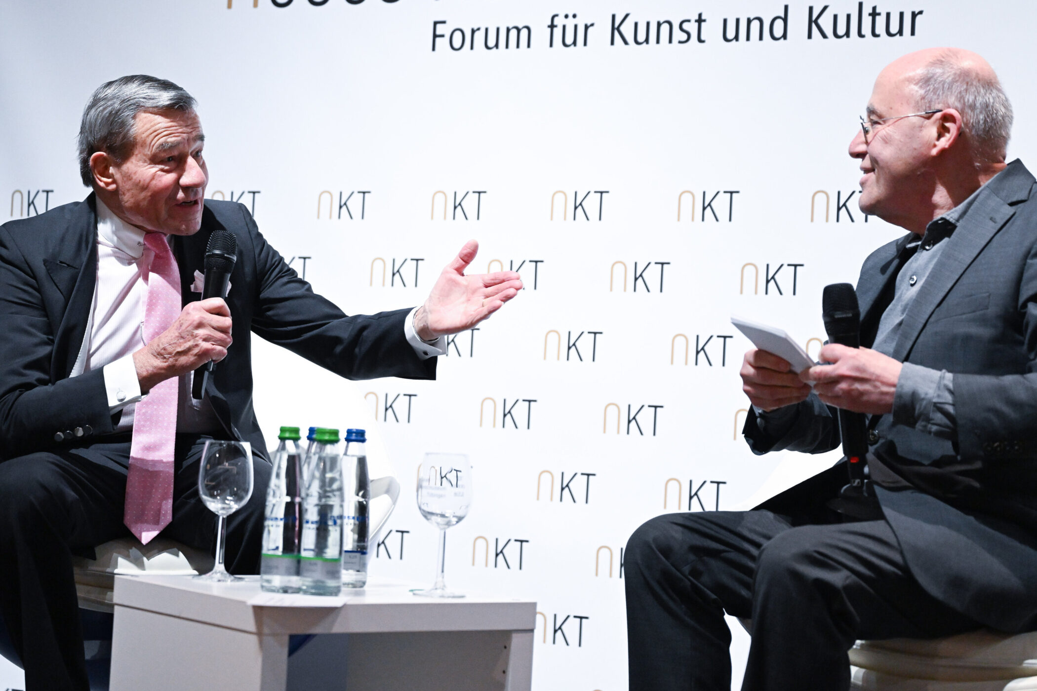 Zwei Männer im Gespräch auf der Bühne mit Mikrofonen. Hintergrund: "Forum für Kunst und Kultur".