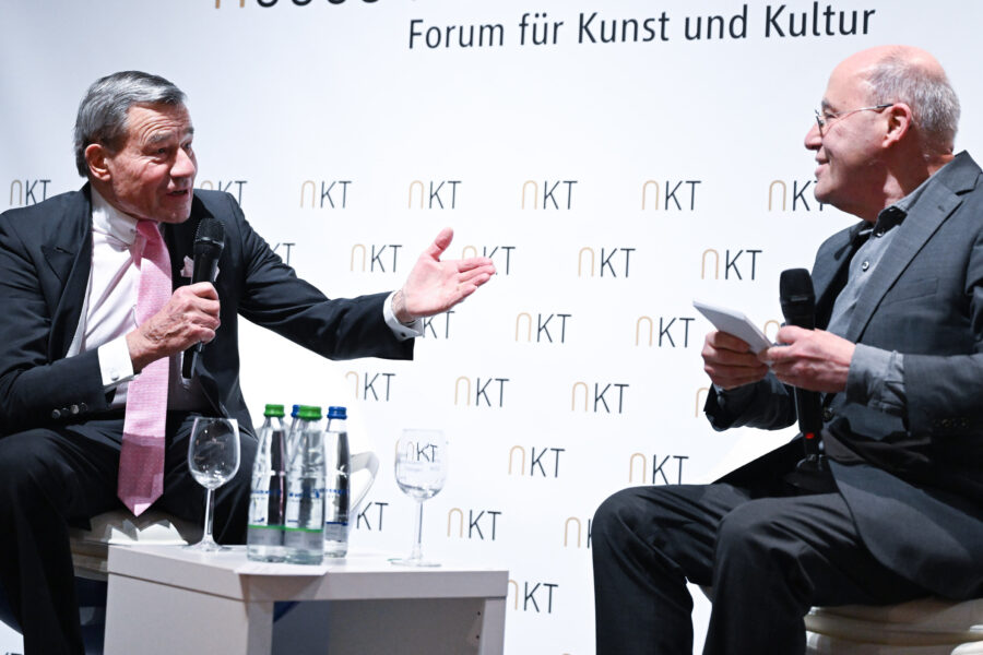 Zwei Männer im Gespräch auf der Bühne mit Mikrofonen. Hintergrund: "Forum für Kunst und Kultur".