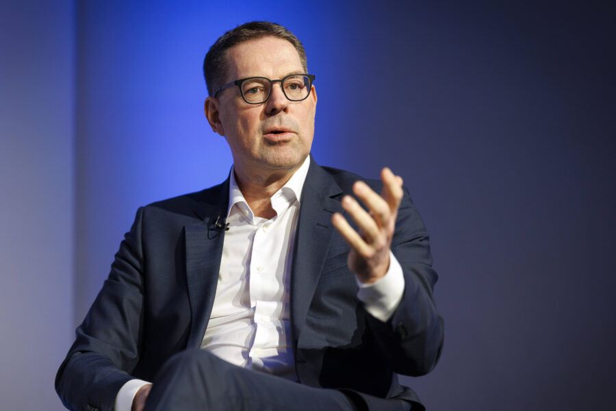 Mann in Anzug spricht gestikulierend vor blauem Hintergrund.