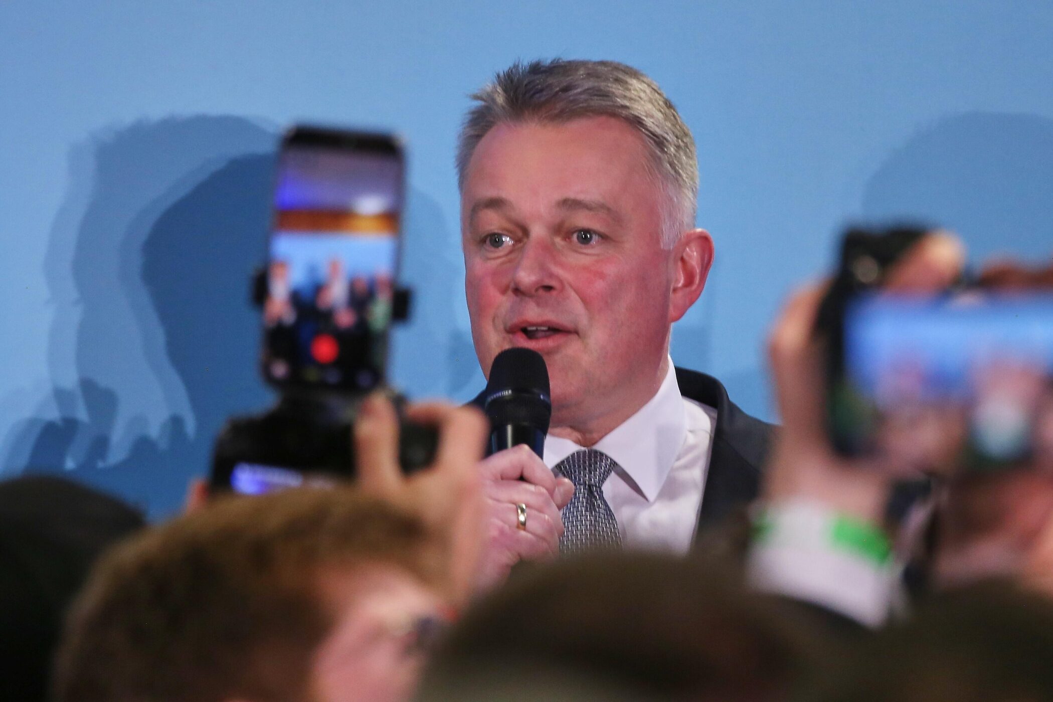 Mann im Anzug spricht in Mikrofon, von Handys umgeben.