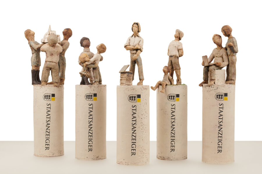 Fünf Skulpturen auf Säulen mit "STAATSANZEIGER"-Schriftzug, einzelne Figuren oben.