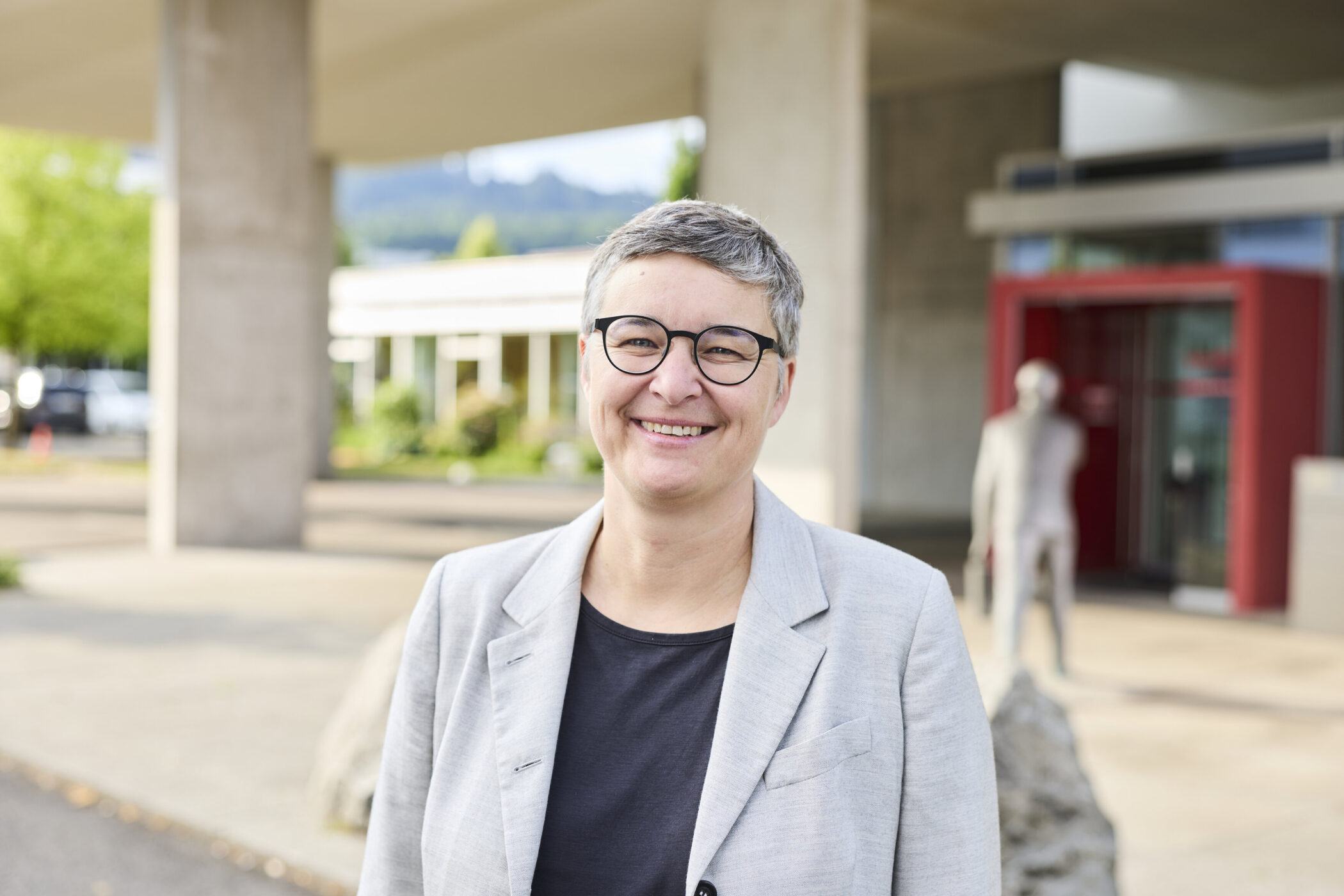 Person mit Brille lächelt in die Kamera, draußen vor einem modernen Gebäude.