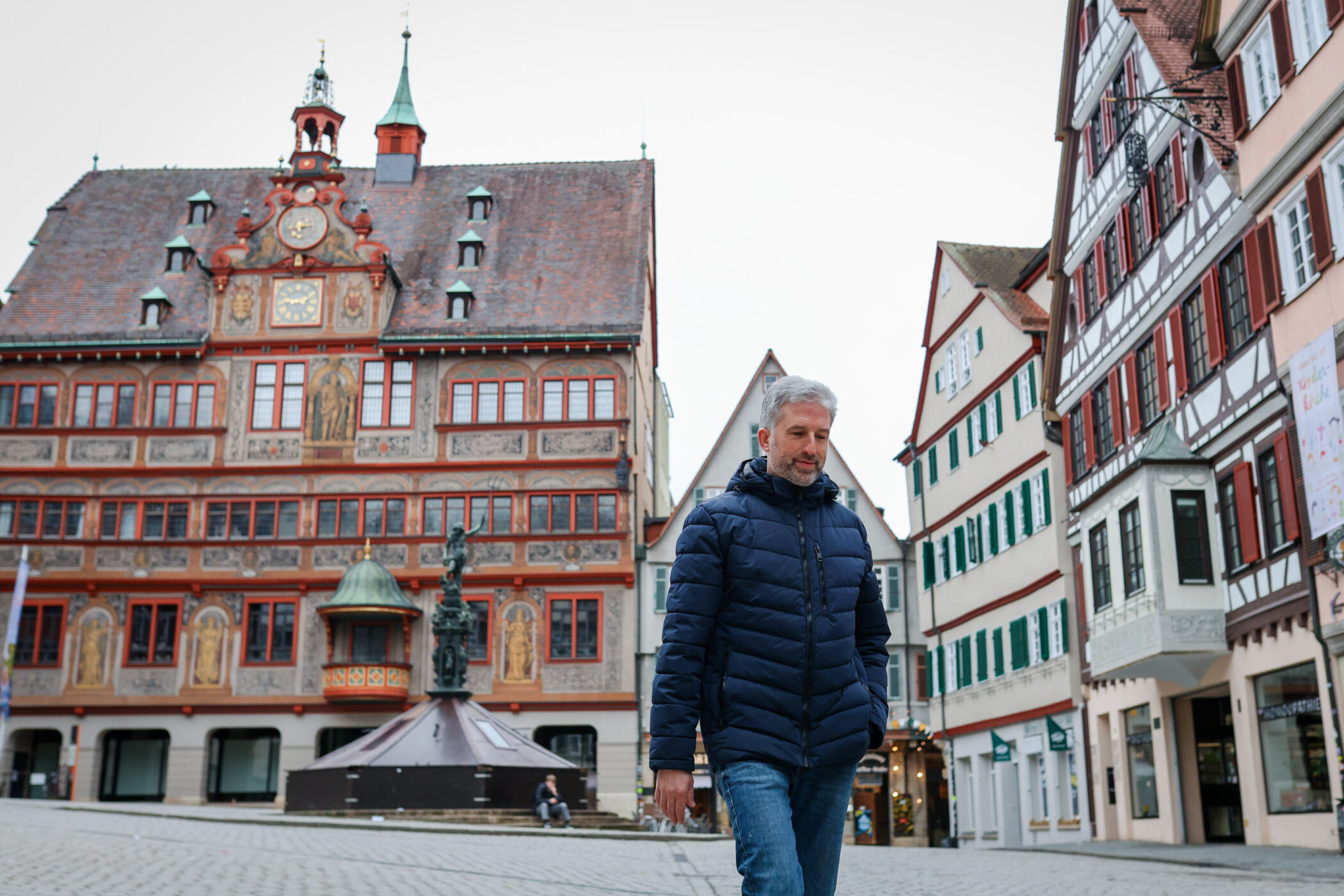 Mann in blauer Jacke geht auf Kopfsteinpflaster, historische Gebäude im Hintergrund.