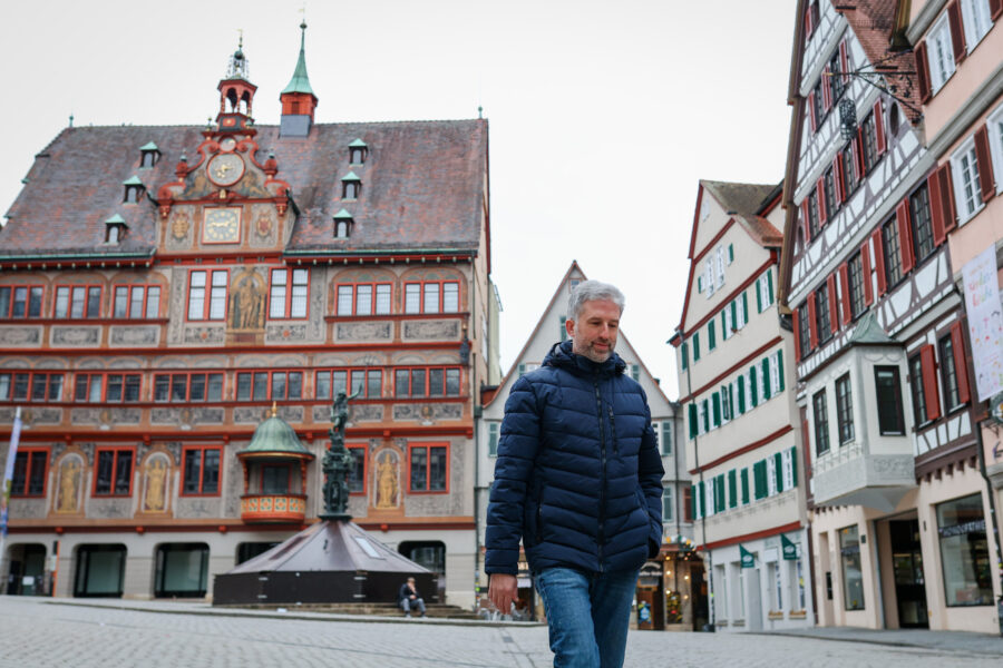 Mann in blauer Jacke geht auf Kopfsteinpflaster, historische Gebäude im Hintergrund.