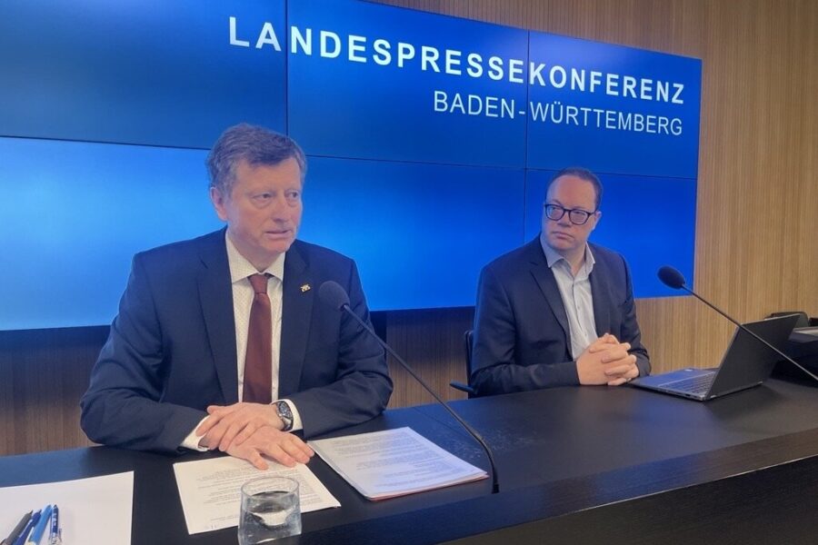 Zwei Männer sitzen bei einer Pressekonferenz, Hinweisschild im Hintergrund.