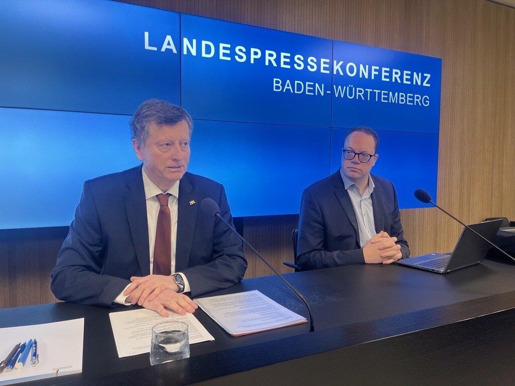 Zwei Männer sitzen bei einer Pressekonferenz, Hinweisschild im Hintergrund.