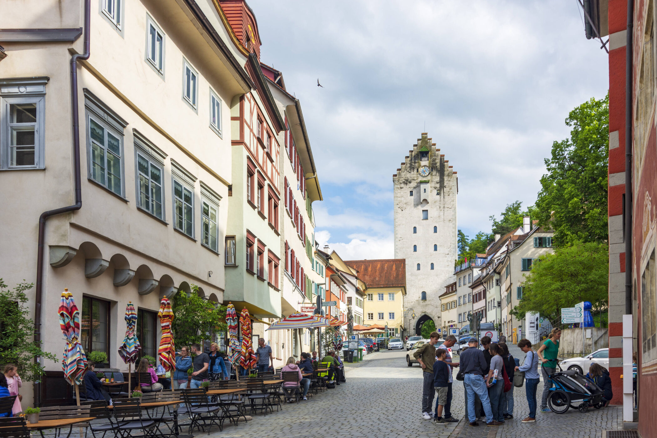 Mehrere Personen in einer gepflasterten Straße vor einem Turm, bunte Sonnenschirme.