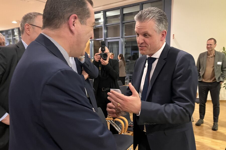 Zwei Männer in Anzügen im Gespräch, Fotograf im Hintergrund.