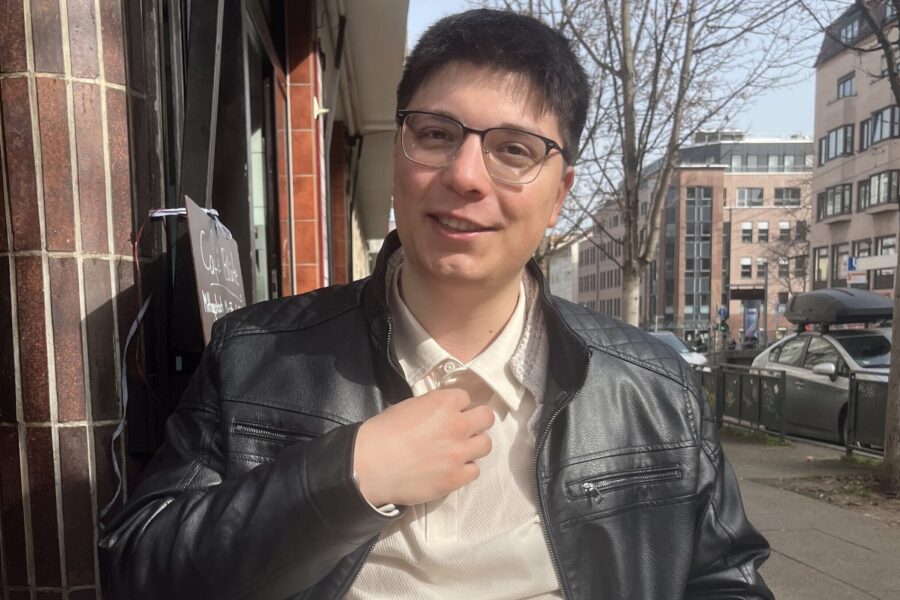 Ein lächelnder Mann mit Brille und Lederjacke steht auf einer Straße.