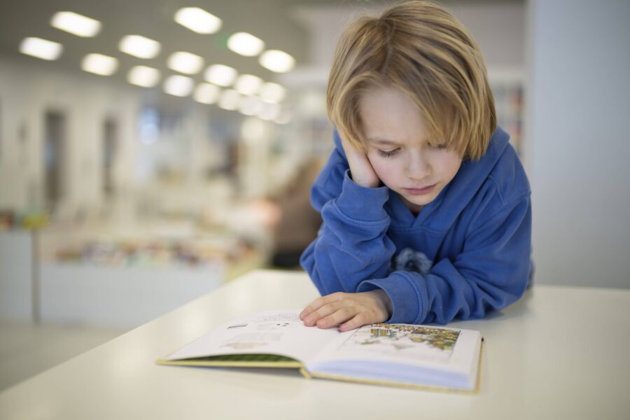 Kind in blauem Hoodie liest Buch an einem Tisch in heller Umgebung.