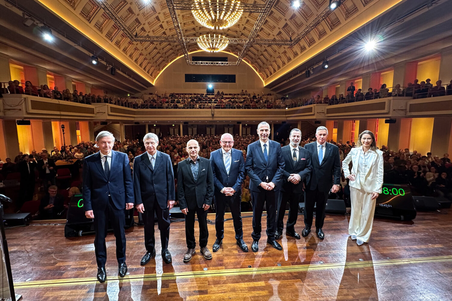 Acht Personen stehen auf einer Bühne in einem großen, vollen Saal.