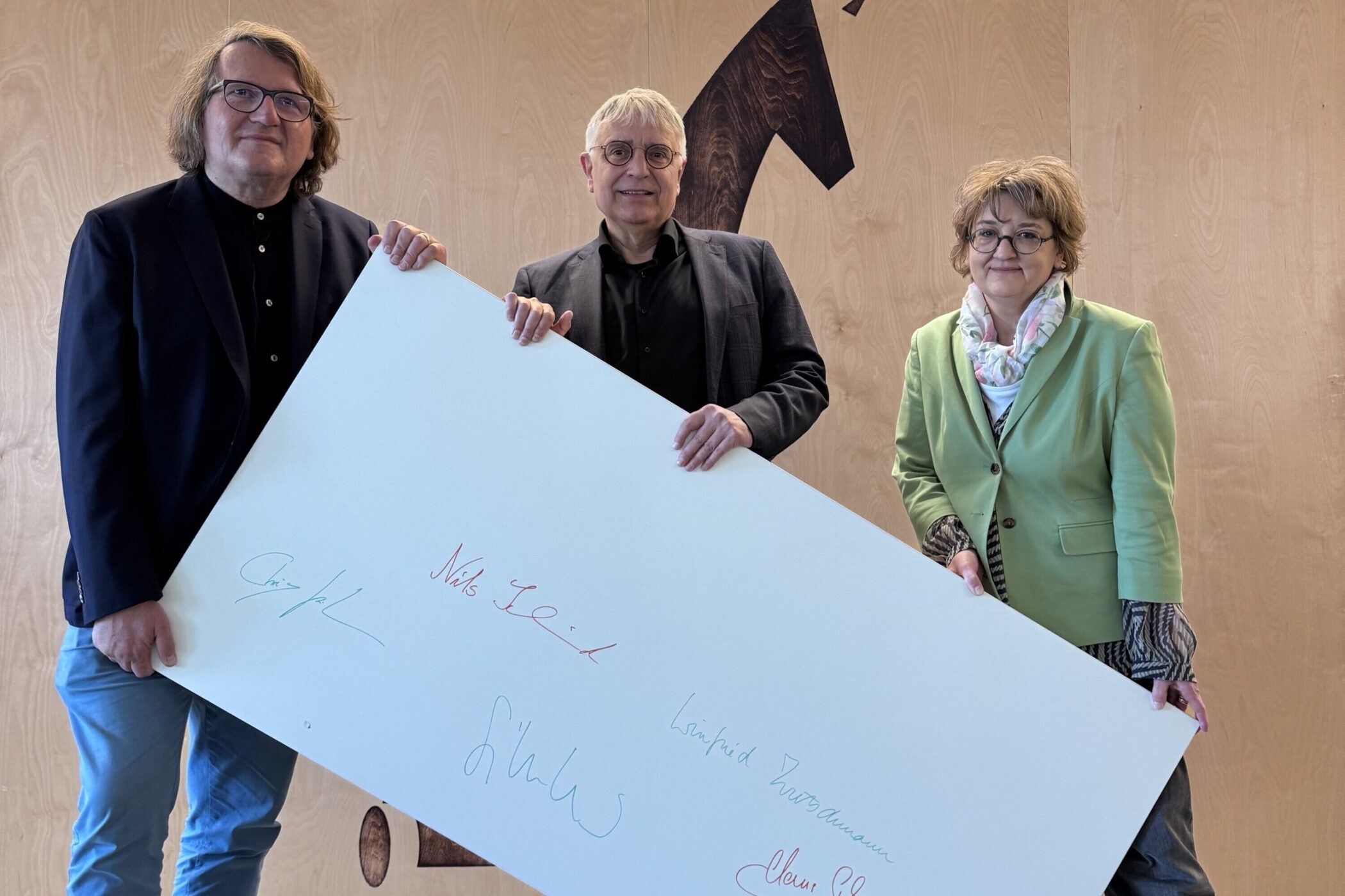 Drei Personen halten großes Brett mit bunten Unterschriften.