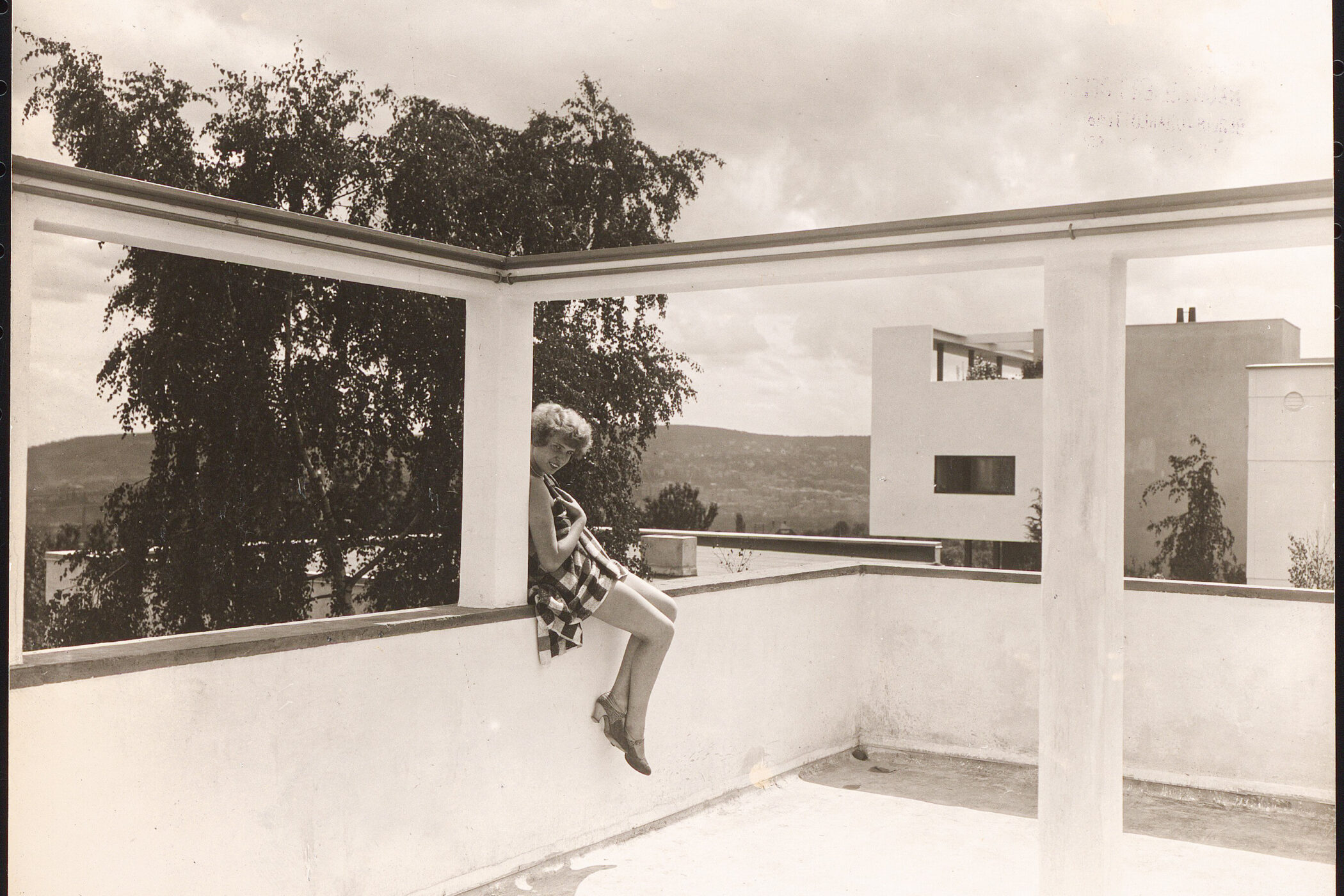 Person sitzt auf Brüstung in modernem Hof, im Hintergrund Bäume und Gebäude.