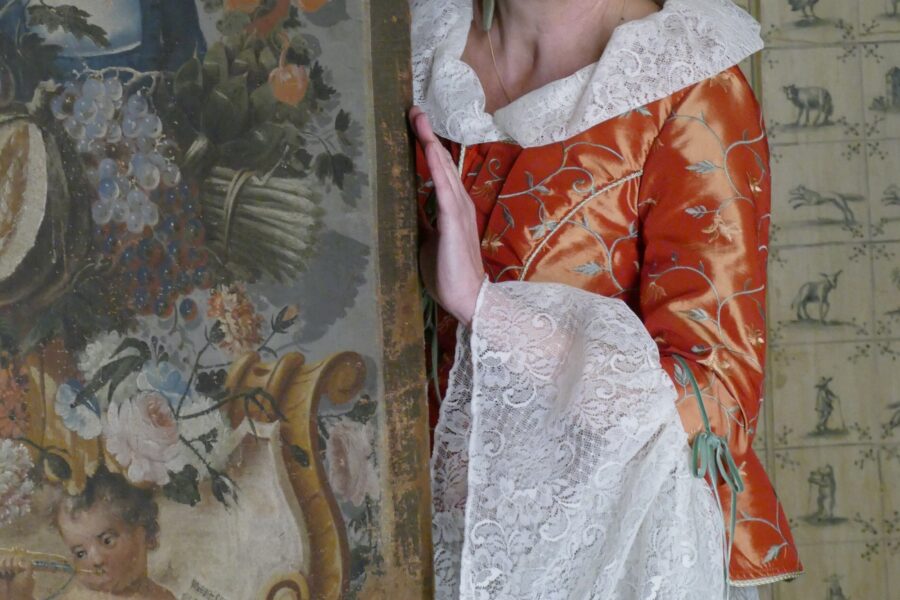 Person in historischer Kleidung neben bemaltem Paneel mit Blumenmotiv.