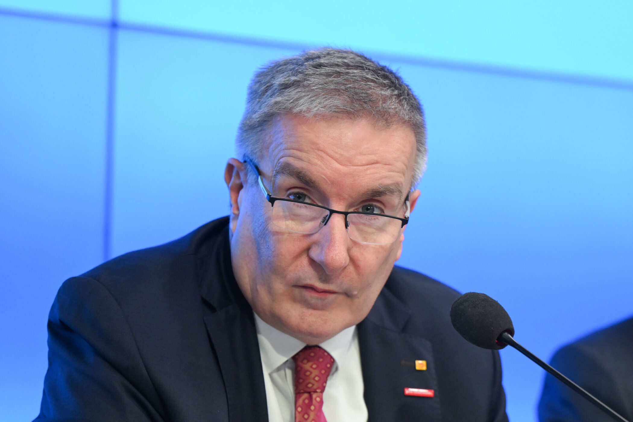 Älterer Mann mit Brille spricht in ein Mikrofon vor blauem Hintergrund.