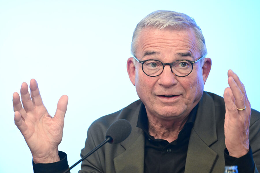 Älterer Mann mit Brille spricht vor Mikrofon, Hände erhoben.