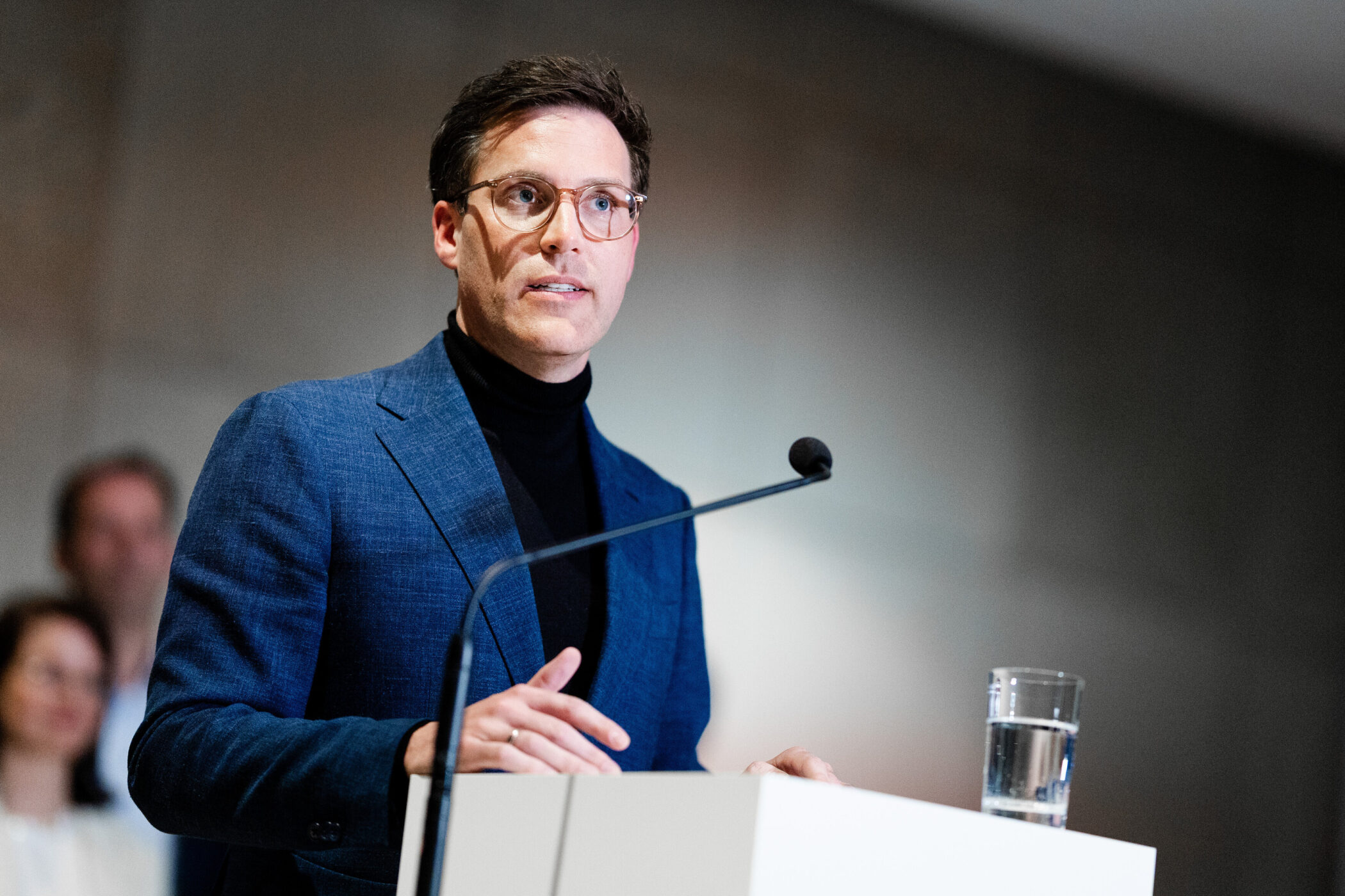 Mann mit Brille und blauem Anzug spricht am Rednerpult, Mikrofon sichtbar.