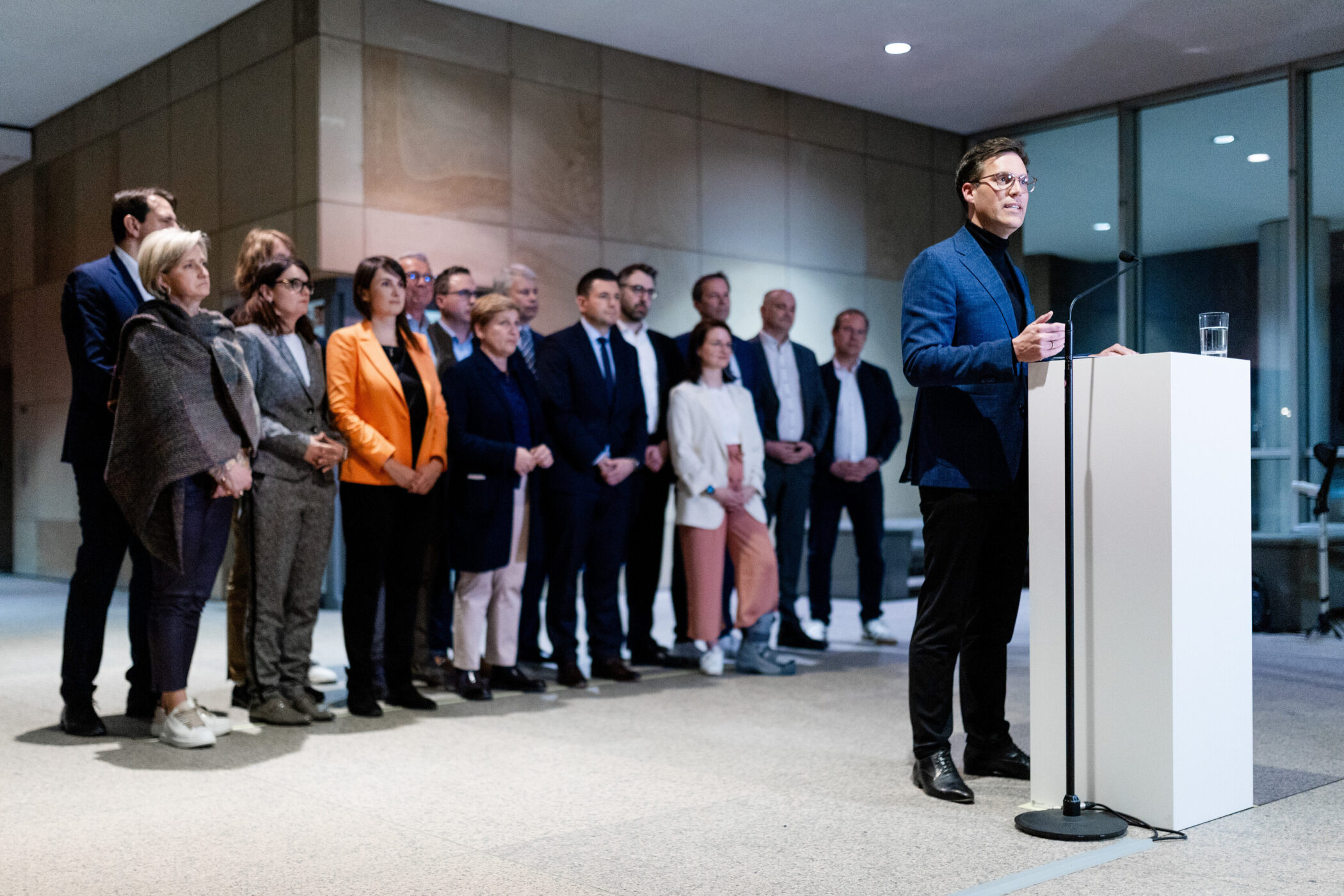 Ein Mann spricht an einem Pult, viele Leute stehen im Hintergrund.