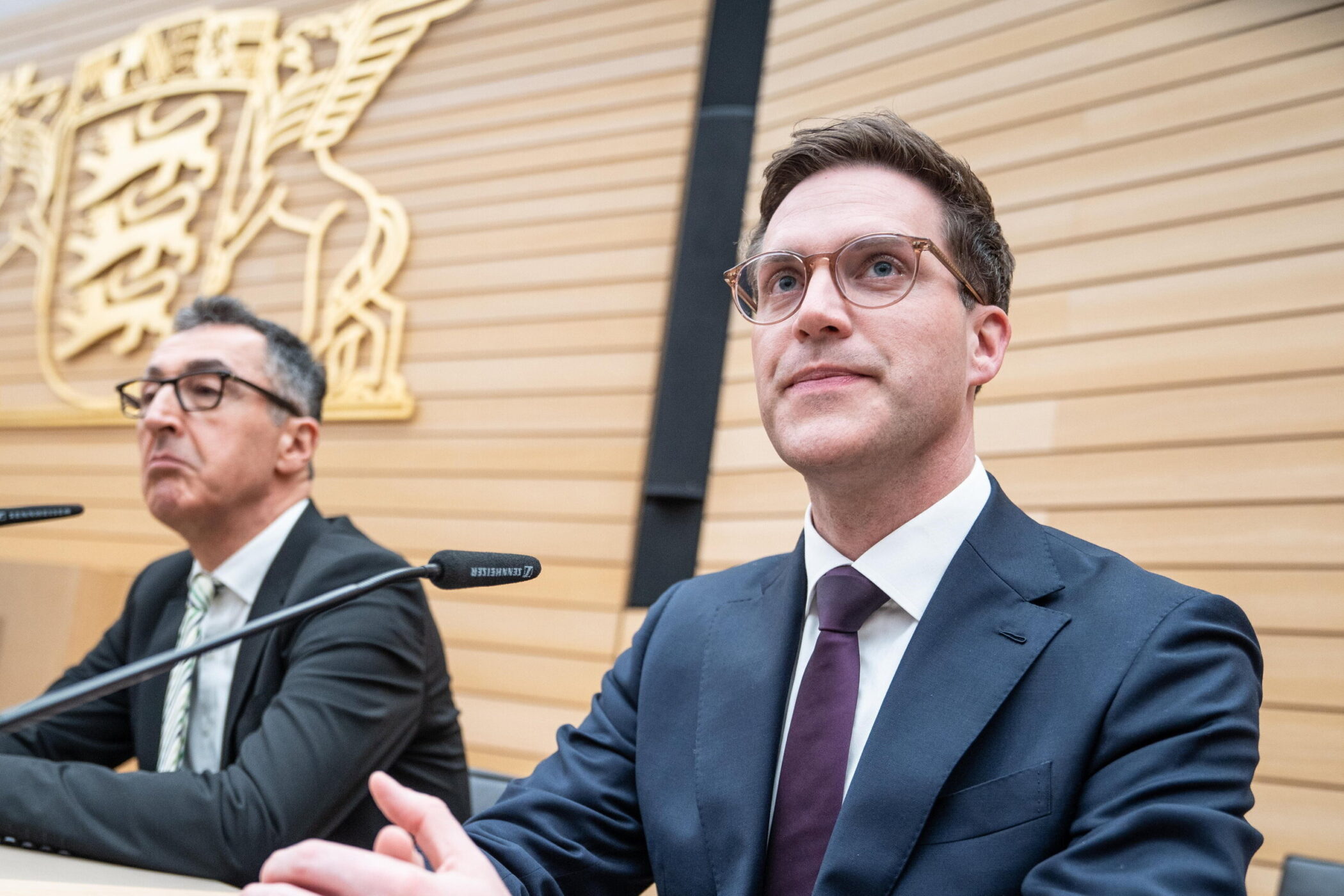 Zwei Männer in Anzügen sitzen an einem Tisch mit Mikrofonen, Holzpaneel im Hintergrund.