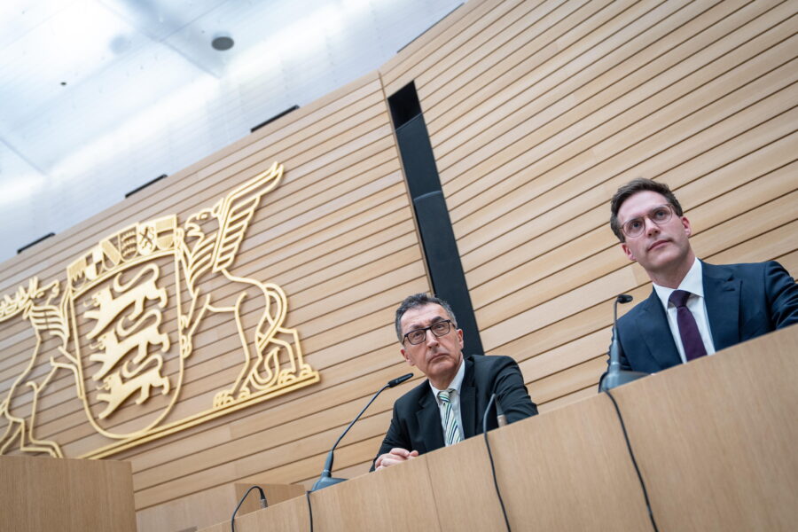 Zwei Männer in Anzügen sitzen an Rednerpulten vor einem großen Wappen.