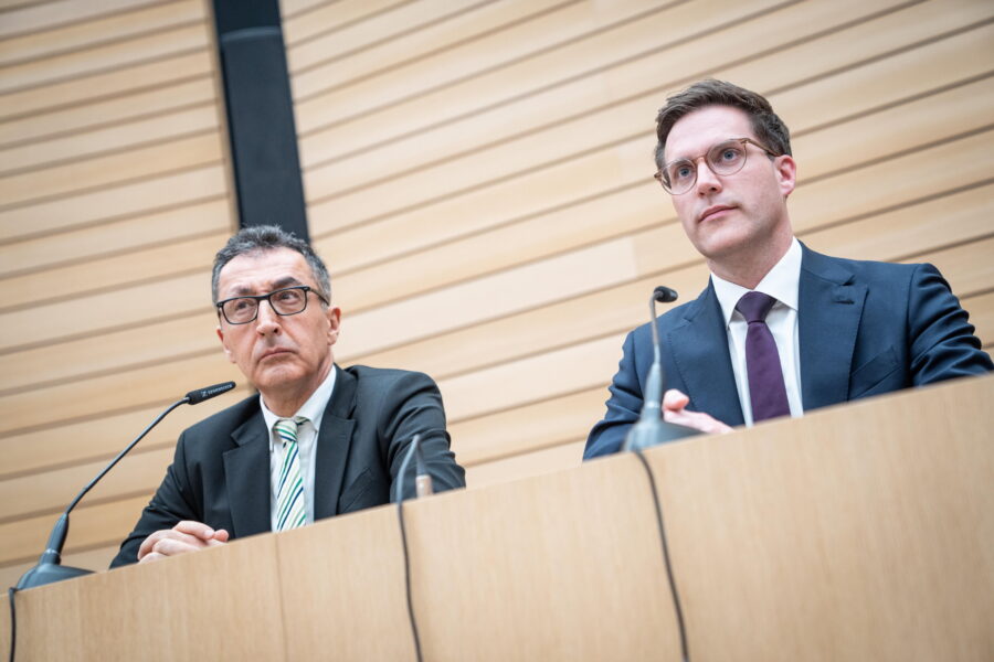 Zwei Männer in Anzügen sitzen hinter einem Tisch mit Mikrofonen.