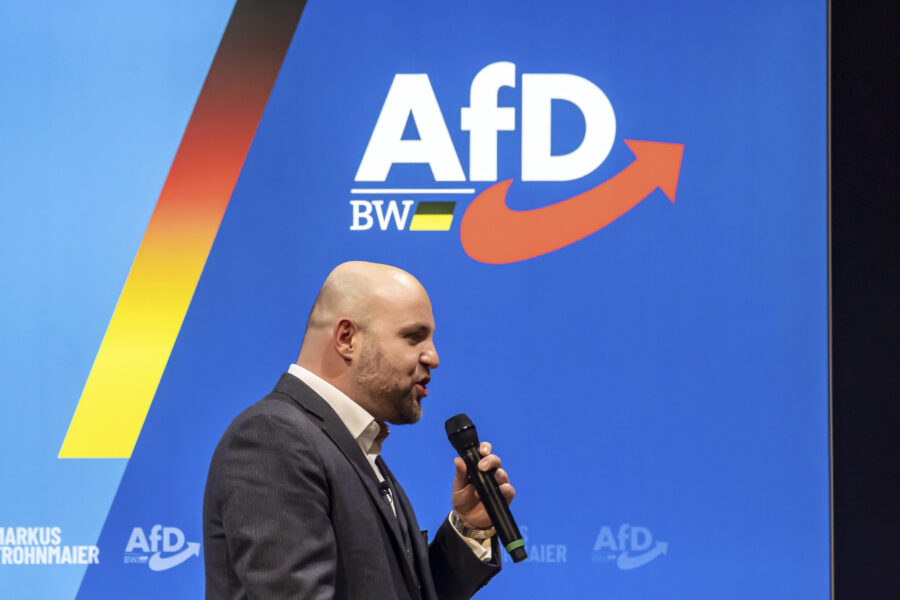 Ein Mann mit Mikrofon vor blauem Hintergrund mit AfD-Logo. Text: Markus Frohnmaier.