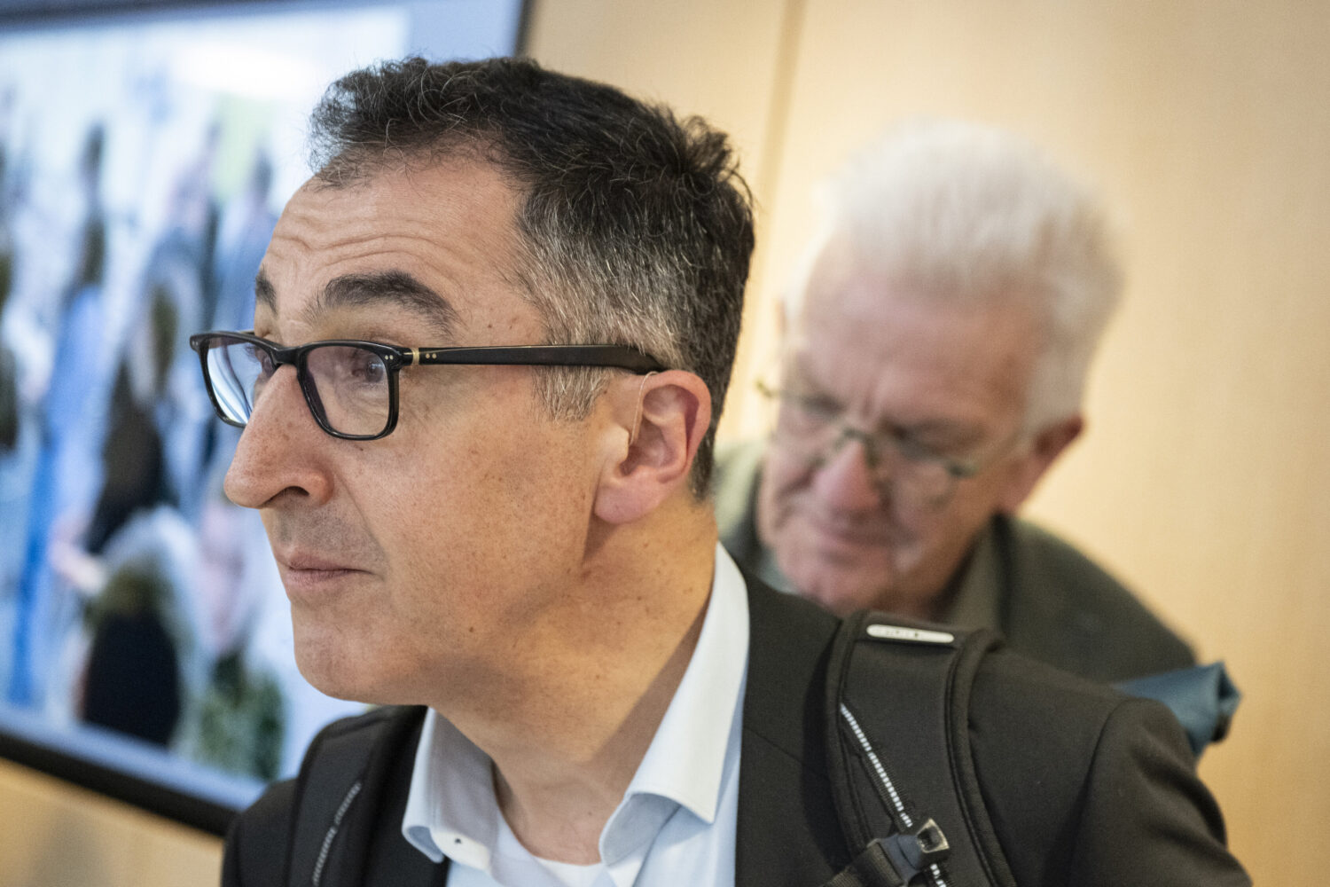 Zwei Männer, einer im Vordergrund mit Brille, ein älterer Mann im Hintergrund.