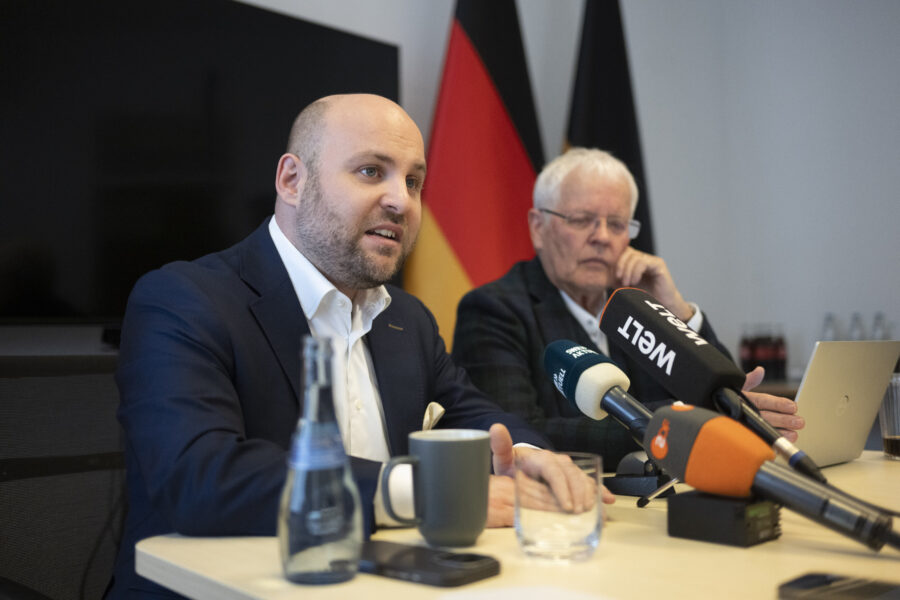 Zwei Männer sitzen, Mikrofone stehen vor ihnen, im Hintergrund eine deutsche Flagge.