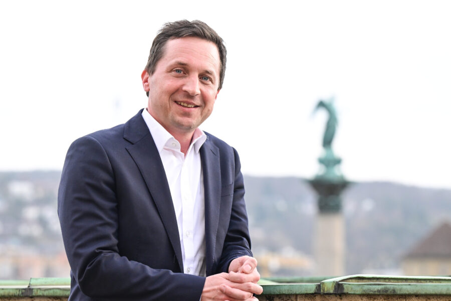 Mann in Anzug, lächelnd, vor unscharfem Hintergrund mit Statue.