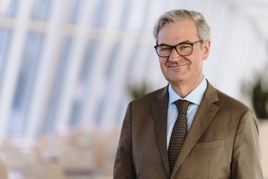 Älterer Mann in Anzug und Krawatte, steht vor unscharfem Hintergrund.