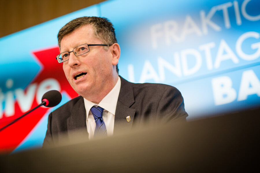 Mann mit Brille am Mikrofon, Hintergrundtext unleserlich.