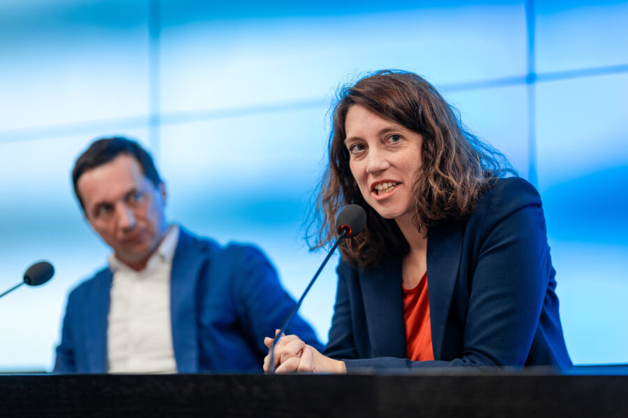 Zwei Personen im Anzug vor Mikrofonen, die Frau spricht. Blauer Hintergrund.