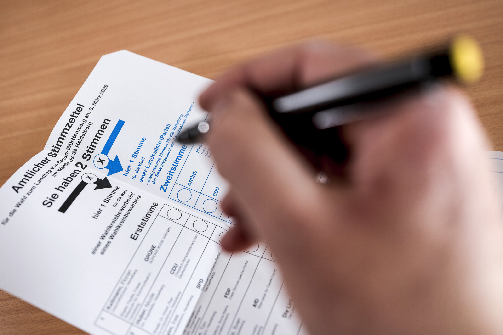 Hand mit Stift über einem ausgefüllten Stimmzettel. Text: "Amtlicher Stimmzettel".