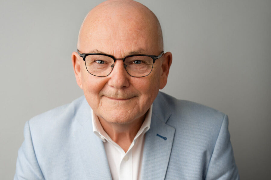 Älterer Mann mit Brille, Glatze, in blauem Sakko vor grauem Hintergrund.