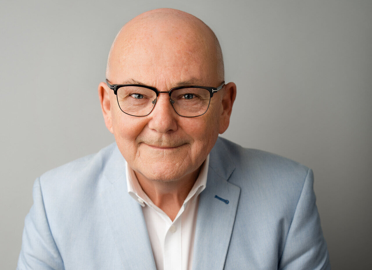 Älterer Mann mit Brille, Glatze, in blauem Sakko vor grauem Hintergrund.