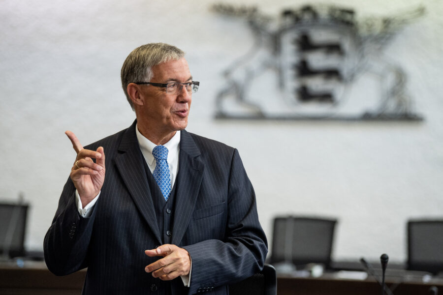 Älterer Mann im Anzug spricht gestikulierend, Büroumgebung im Hintergrund.