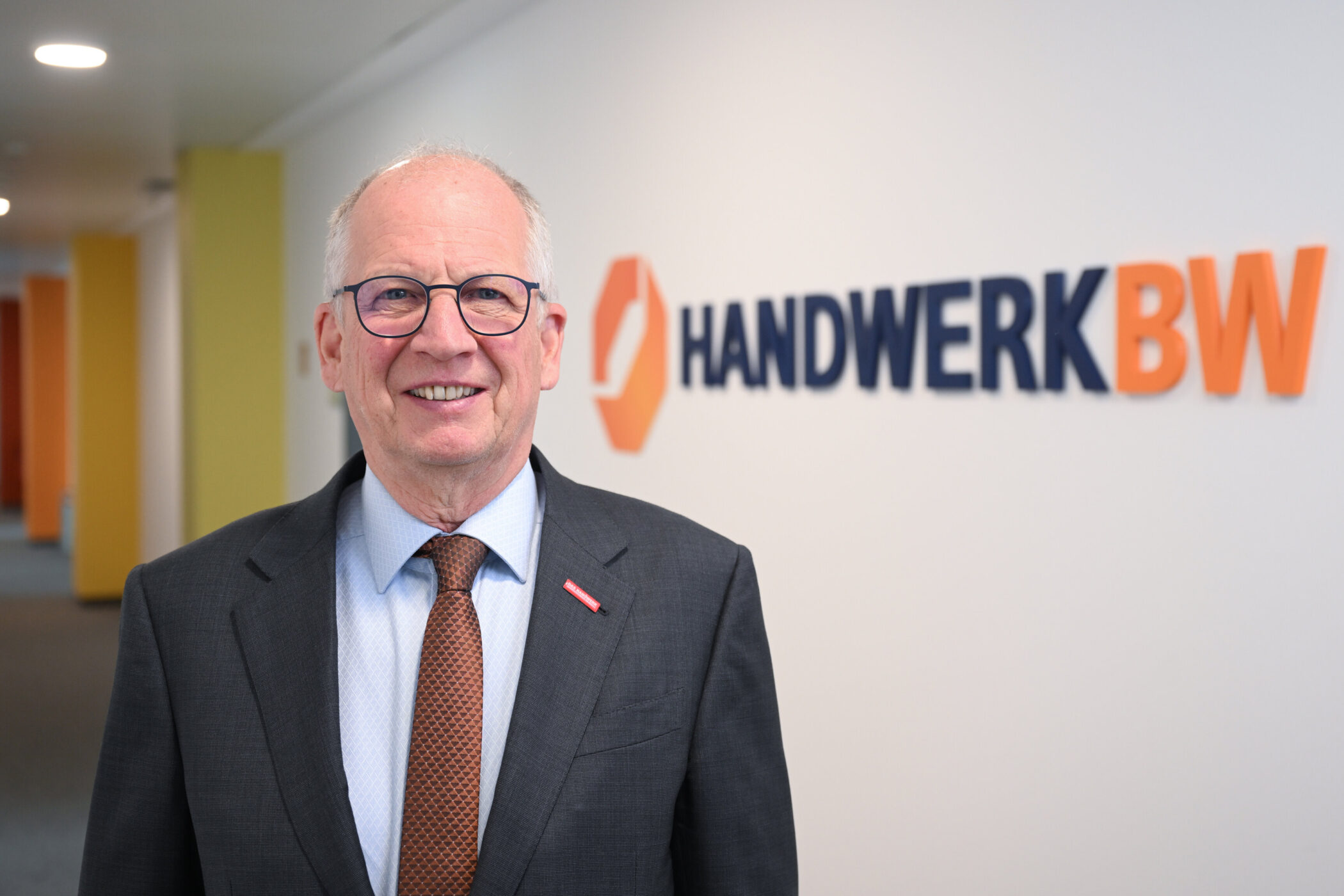 Älterer Mann mit Brille in Anzug vor "HANDWERK BW" Logo an Wand.