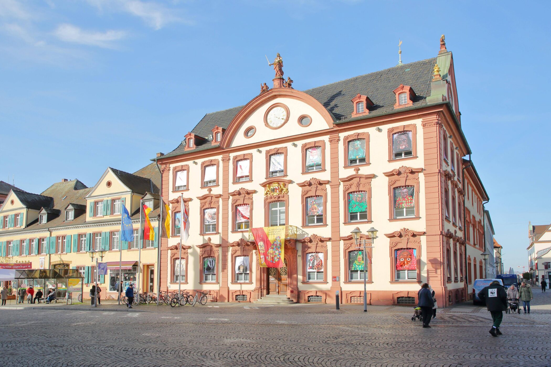 Mehrstöckiges Gebäude mit verzierten Fenstern, Menschen auf Kopfsteinpflasterplatz.
