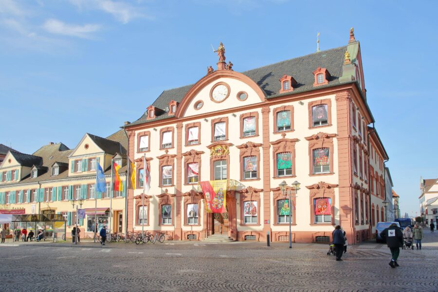Mehrstöckiges Gebäude mit verzierten Fenstern, Menschen auf Kopfsteinpflasterplatz.