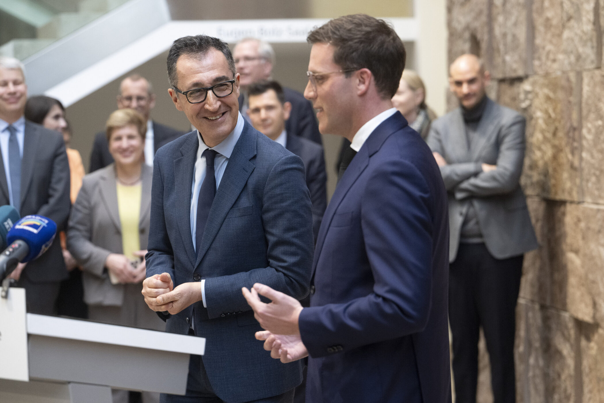 Zwei Männer in Anzügen im Gespräch, umgeben von mehreren Personen.