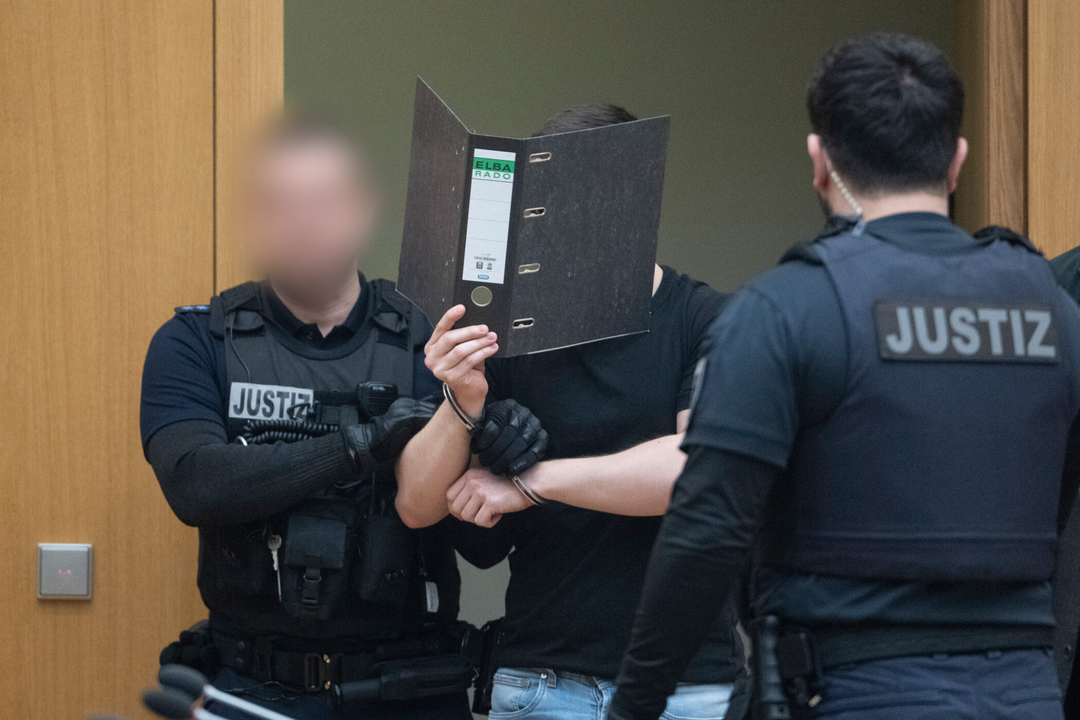 Drei Personen in schwarzer Justizkleidung, eine mit Aktenordner im Gesicht.