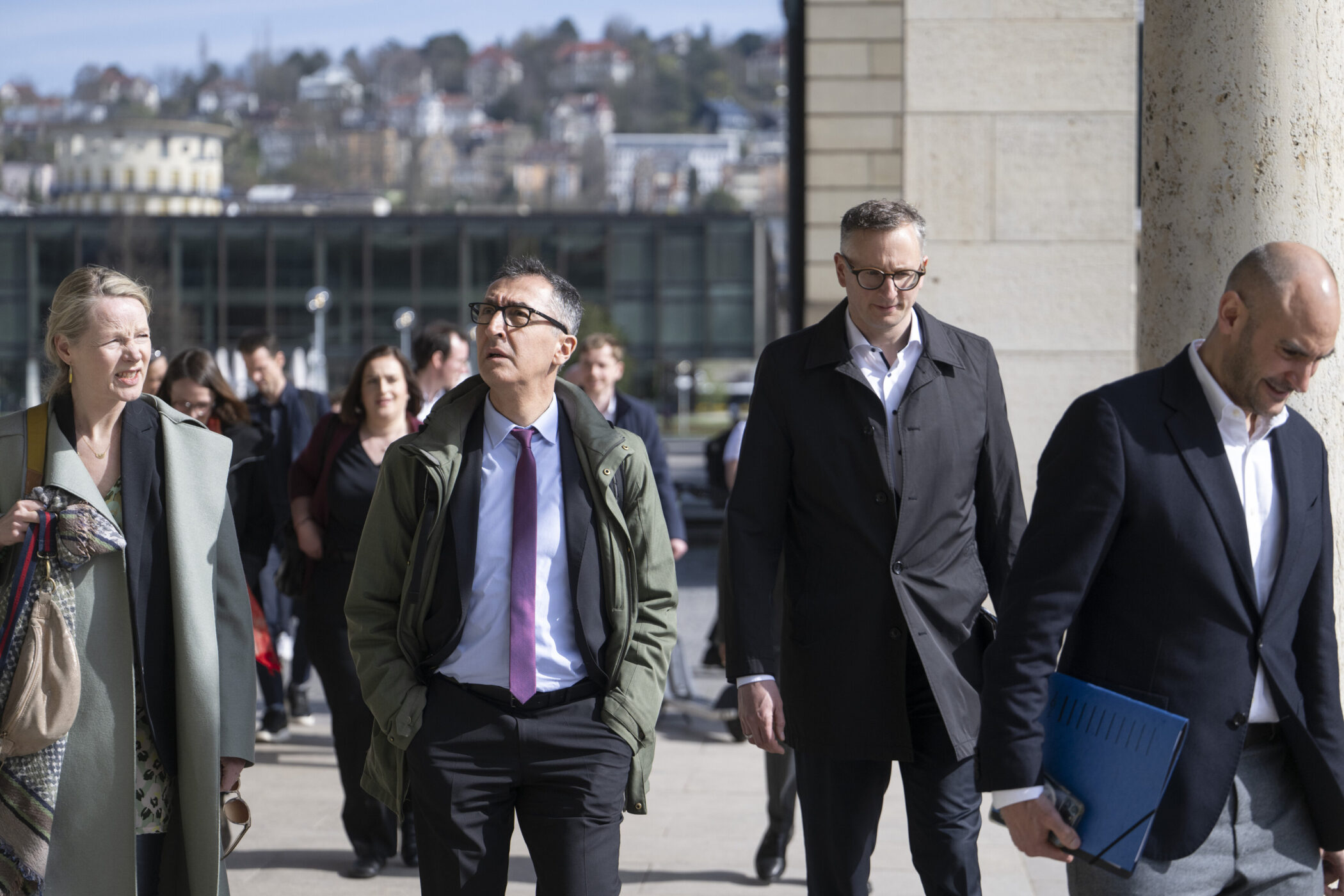 Mehrere Personen in Anzügen auf Gehweg, Gebäude und Bäume im Hintergrund.