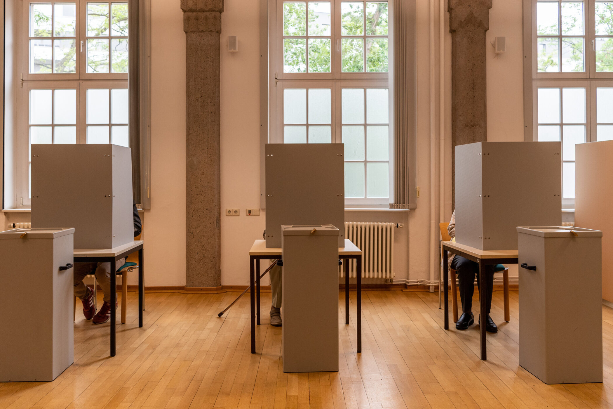 Menschen wählen in Wahlkabinen in einem hellen Raum mit Holzboden.