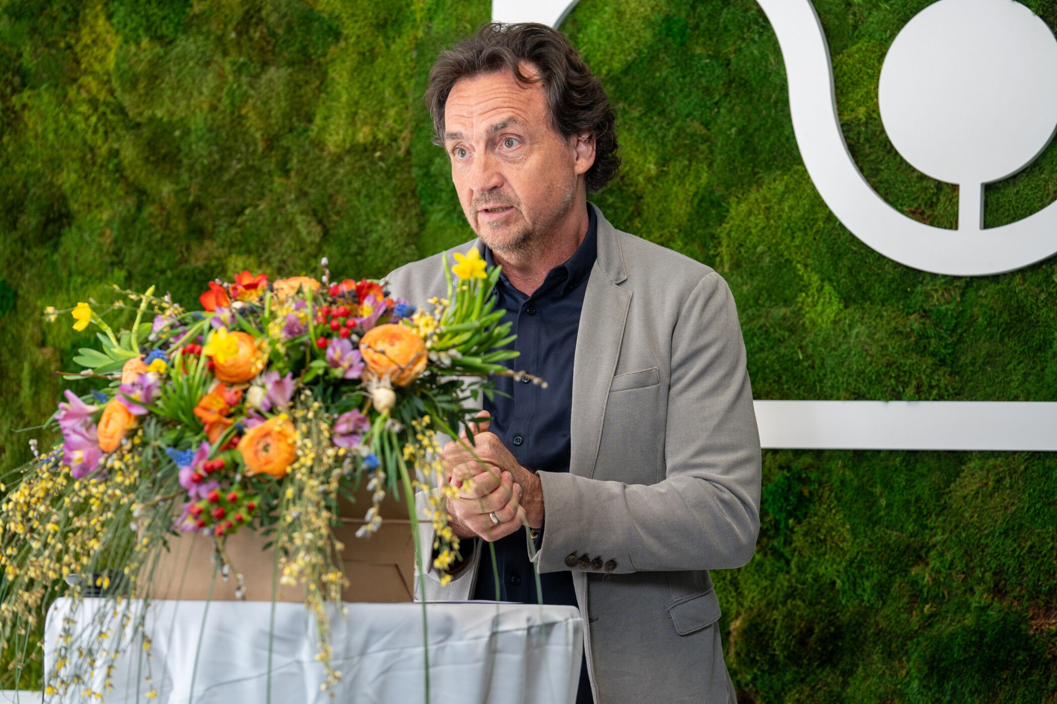 Mann im Anzug spricht hinter Blumenstrauß, grüner Hintergrund.