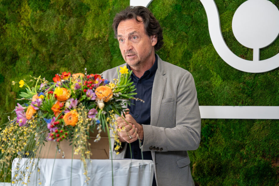 Mann im Anzug spricht hinter Blumenstrauß, grüner Hintergrund.