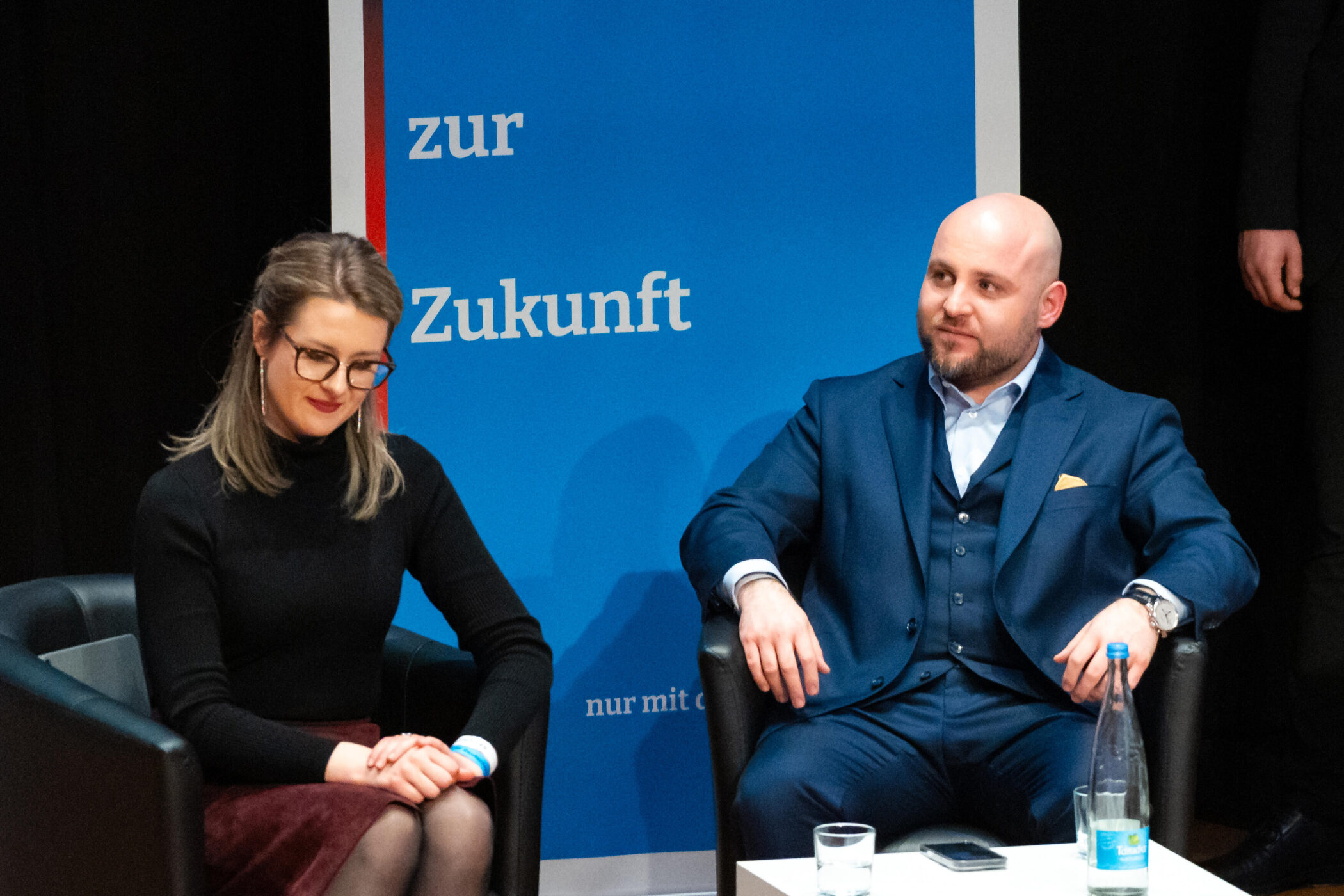 Zwei Personen sitzen nebeneinander, hinter ihnen ein blaues Schild mit "zur Zukunft".