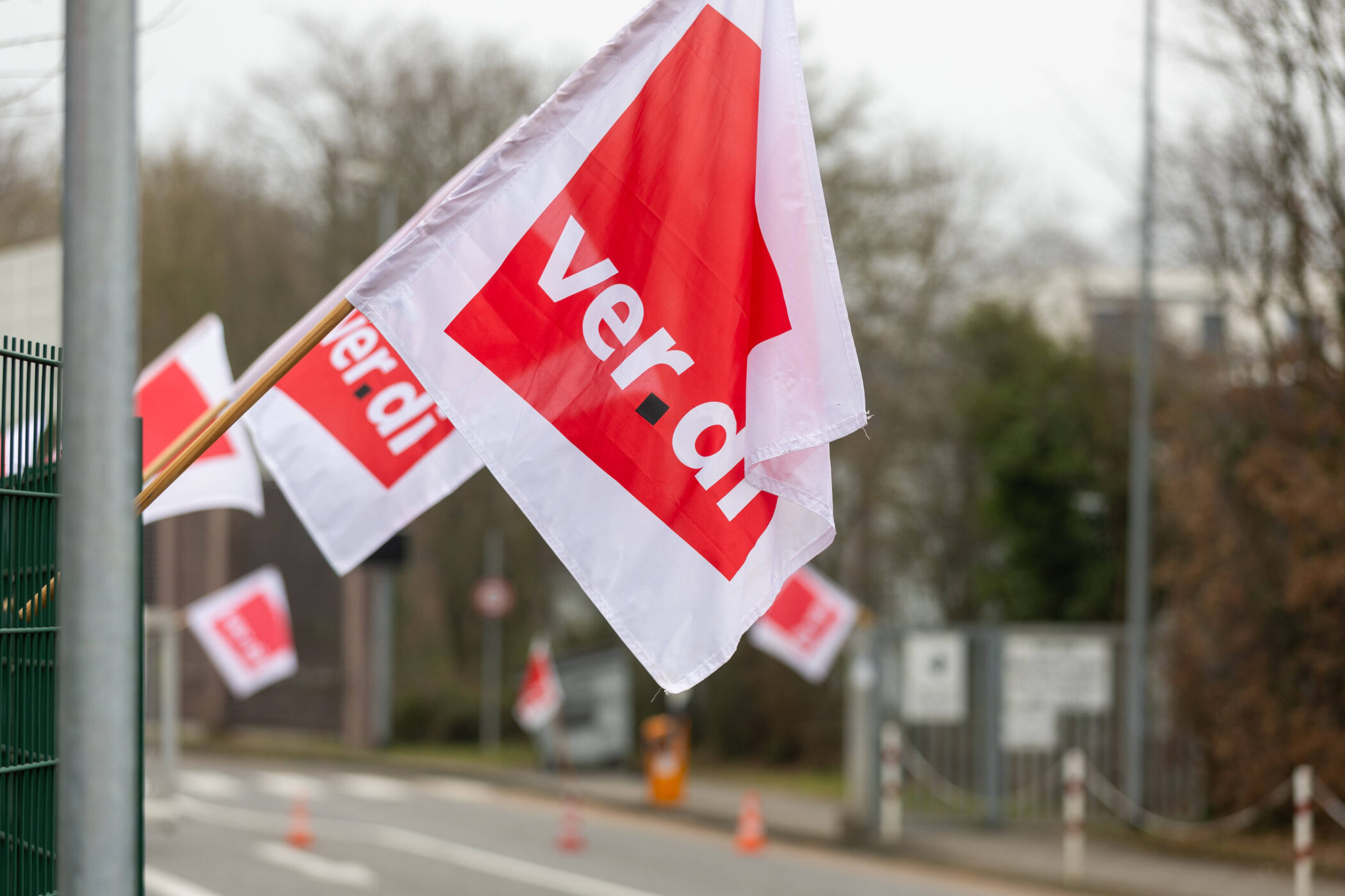 Weiße Fahne mit rotem Logo und Text "ver.di" weht im Freien an einem Zaun.