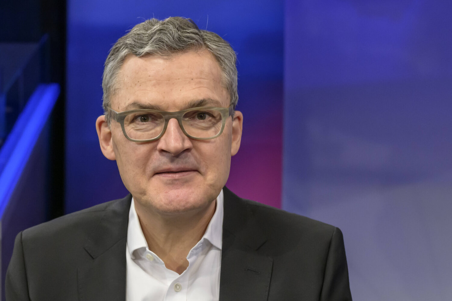 Eine Person trägt Brille, Anzug und Hemd, vor blauem Hintergrund.